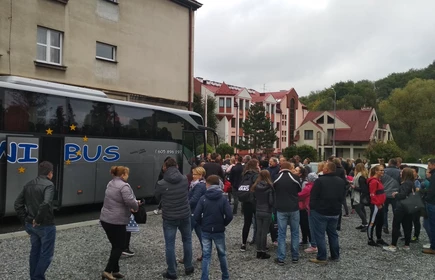 Na zdjęciu grupa os&oacute;b zbierająca się na parkingu na tle autobusu